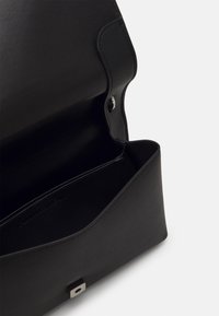 Černá kožená kabelka s hladkou texturou, ostrými tvary a patentním uzávěrem. Interiér se vyznačuje minimalistickým designem a detaily šití.