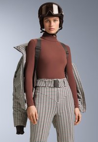 Kvinna i brun polotröja, rutiga högmidjade byxor med bälte, håller en matchande jacka, bär svart skid hjälm och skidglasögon.