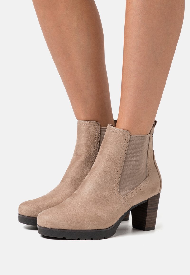 Bottes beige en daim montant à la cheville, à talons carrés, avec panneaux élastiques sur les côtés, portées sur des jambes nues sur un fond neutre.
