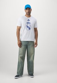 Hvid bomulds T-shirt med blåt Real Madrid-logo, parret med lyseblå denim bukser med bred ben og blå kasket. Skoene er marineblå med hvide såler.