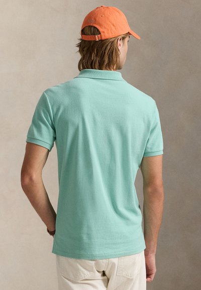 Mies, jolla on mintunvihreä poolopaita, beiget housut ja oranssi lippalakki, kasvot poispäin neutraalia taustaa vasten.