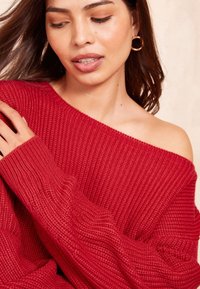 Pulover roșu tricotat cu design off-shoulder, textură groasă și mâneci largi. Țesătura pare moale și are un stil casual.