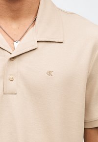 Μπεζ πολο με υφή, δύο κουμπιά και λογότυπο "cK" στο στήθος, φορεμένο με ασημένιο ορθογώνιο μενταγιόν σε αλυσίδα.