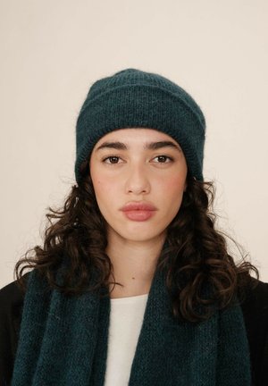 Bonnet en tricot turquoise avec un tissu texturé et un revers replié, associé à une écharpe assortie, porté sur une chemise blanche. Fini lisse et chaleur.