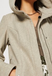 Giacca grigia in misto lana con cappuccio, dotata di una cerniera frontale, tasche laterali e una texture liscia. Accentata da un top marrone sotto.