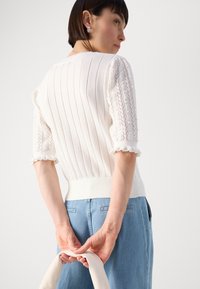 Maglione bianco a maglia con texture a coste e maniche in pizzo, abbinato a jeans blu chiaro. Presenta una vita definita e bordi delle maniche arricciati.