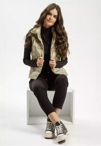 Jasno beżowa kamizelka na zamek, założona na czarny golf, w zestawieniu z czarnymi legginsami oraz czarno-białymi sneakersami typu high-top, siedząca na sześcianie.