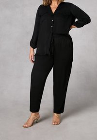 Svart, löst sittande silkestopp med två framfickor, kombinerad med svarta, avsmalnande byxor med sidofickor och dragsko i midjan. Guldfärgade klackskor.