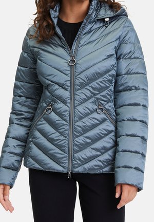 Femme portant une doudoune matelassée bleu métallique avec coutures diagonales, fermeture éclair à l'avant et poches, ainsi qu'une capuche sur un top noir.