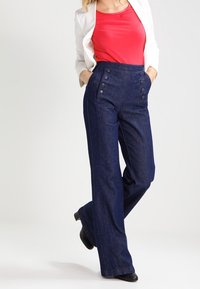 Marineblå denim bukser med bred ben og høj talje, med sidelommer og seks store knapper foran, kombineret med en rød top og en hvid blazer.