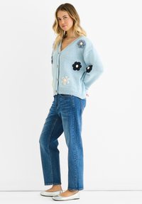 Light blue knitted cardigan with floral appliqué embellishments, v-neck, and button closure, paired with straight-leg blue jeans and silver flats.
