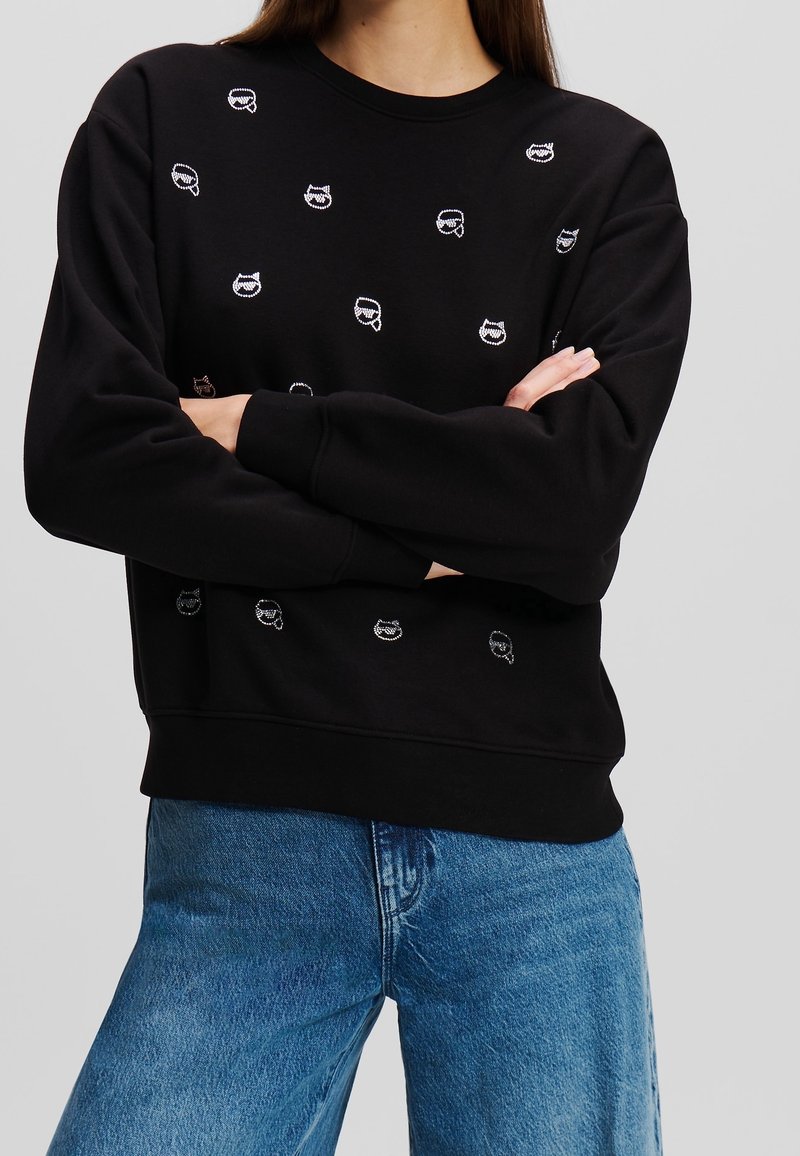 Personne portant un sweat-shirt noir avec de petits motifs de visages de chouettes blanches, bras croisés, associé à un jean bleu en denim.