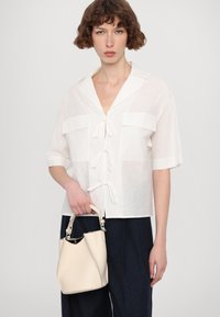 Witte korteblaussjes met een kraag, knoopsluiting aan de voorkant en twee borstzakken. Houdt een kleine, crèmekleurige leren handtas vast met een gebogen handvat.