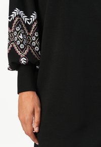 Zwarte jurk met een patronenmouw met bloemen- en diamantontwerpen in wit, roze en groen. Elastische manchetten zorgen voor structuur.