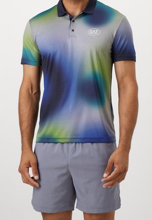 Mann trägt ein kurzärmeliges Polohemd mit blau-grünem Farbverlauf und EA7-Logo auf der Brust und hellgraue Shorts.