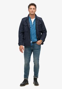 Mørkeblå jakke med flere lommer, lagd over en lyseblå skjorte. Båret med slim-fit jeans og sorte snørebåndsstøvler.