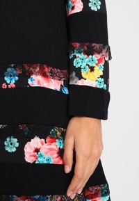 Svart klänning i tyg med blommönster i röd, blå och gul. Utsmyckad med spetsdetaljer och olika blommönster på ärmarna.