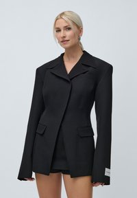 Femeie blondă îmbrăcată într-un sacou negru oversized cu umeri accentuați și mâneci lungi, asociat cu pantaloni scurți negri, pozând pe un fundal alb.