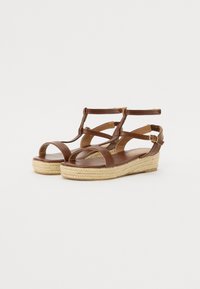 Brown leather wedge sandals with a woven jute sole, featuring crisscross straps and ankle buckle. Smooth texture, open toe design.
