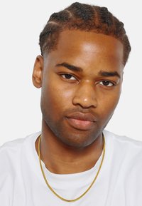 Jeune homme avec des cheveux tressés portant une chemise blanche et un collier en chaîne dorée. Peau lisse et expression neutre, regardant droit devant.
