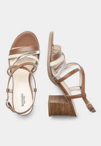 Sandales à talons en cuir marron avec des brides croisées, des accents dorés et un talon bloc strié. La semelle intérieure est beige clair avec un logo.