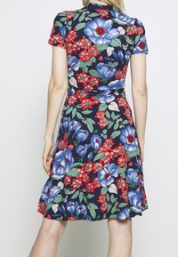 Donna che indossa un vestito blu navy al ginocchio con grandi motivi floreali blu e rossi, vista da dietro su uno sfondo semplice.