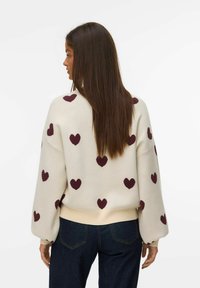 Femme aux longs cheveux bruns portant un pull crème avec des motifs de cœurs violets et un jean bleu foncé, de dos contre un fond uni.