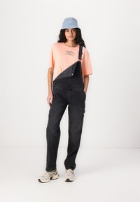Zwarte denim overall met één schouderband, een roze graphic t-shirt eronder en een lichtblauwe bucket hat. Sneakers maken de outfit compleet.