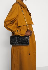 Borsa a tracolla nera in pelle con una texture a coccodrillo, forma rettangolare e tracolla in catena dorata, indossata sopra un cappotto giallo senape.