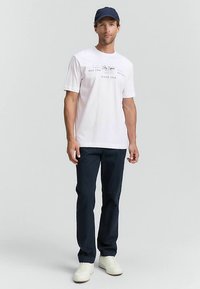 Weißes T-Shirt mit aufgedrucktem Text, kombiniert mit dunkelblauen Hosen. Model trägt eine blaue Kappe und weiße Schuhe, steht vor einem neutralen Hintergrund.