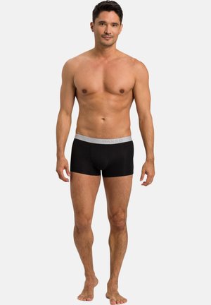 Mann trägt schwarze Boxer-Shorts mit grauem Bund, steht barfuß vor hellem, einfarbigem Hintergrund.