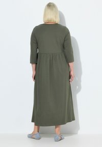 Sukienka maxi w oliwkowym kolorze z bawełny, z okrągłym dekoltem, rękawami o długości trzech ćwiartek, marszczonym pasem i gładką teksturą. Połączona z jasnoniebieskimi butami.