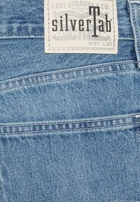 Jeans de denim azul claro con una etiqueta blanca que dice "silverTab" e incluye el tamaño W31 L32. La tela tiene un tejido texturizado.