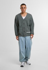 Siv krožnik s V-izrezom, gumbi spredaj, dvema žepoma in podrobnostjo z besedilom "drugo". Uklopljen s svetlo modrimi širokimi džinsami in športnimi copati.