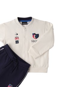 Chaqueta blanca con cremallera y acentos en azul marino, combinada con pantalones de cintura elástica en azul marino que tienen rayas laterales.