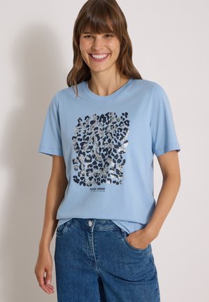 Femme souriante portant un t-shirt bleu clair avec un imprimé léopard sombre et argenté et un jean bleu, debout devant un fond clair uni.