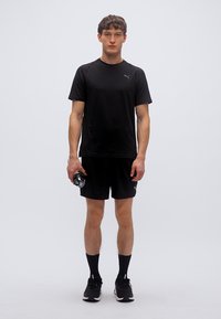 Μαύρο αθλητικό μπλουζάκι και σορτς από ύφασμα που απομακρύνει την υγρασία. Το μπλουζάκι έχει κοντά μανίκια και ένα διακριτικό λογότυπο. Το μοντέλο κρατάει ένα μπουκάλι νερό.