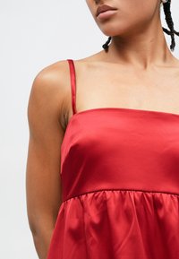 Rochie din satin roșu, cu bretele subțiri, corsaj strâmt și fustă evazată cu detalii drapate în jurul taliei. Textură fină și finisaj lucios.