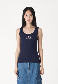 Top din bumbac ribbed de culoare albastru închis, cu o croială cu decolteu rotund și logo-ul alb "GAP" pe față. Asortat cu o fustă din denim cu buzunare.
