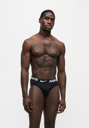 Muž s vyrysovanou postavou stojí pred jednoliatym pozadím a má na sebe čierne Nike slipy s viditeľným logom na páse.