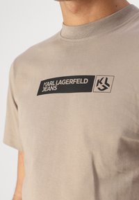Bela bombažna T-majica z dolgimi rokavi z črnim pravokotnim logotipom z napisom "KARL LAGERFELD JEANS" in stiliziranim grbom na levem prsnem predelu.