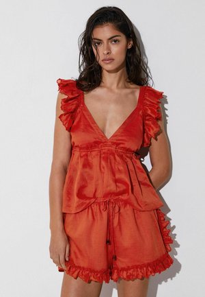 Naine seisab näoga ettepoole, kandes punast varrukateta topi volditud detailidega ja sobivaid lühikesi pükse ühevärvilise tausta ees.