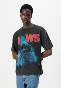Svart grafisk t-skjorte med et stort blåtig haidesign og røde "JAWS"-tekst. Korte ermer og avslappet passform, med en myk tekstur.