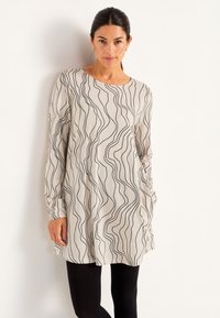 Vrouw gekleed in een beige tuniek met lange mouwen en zwarte golvende lijnen, gecombineerd met zwarte leggings, staand tegen een witte muur.