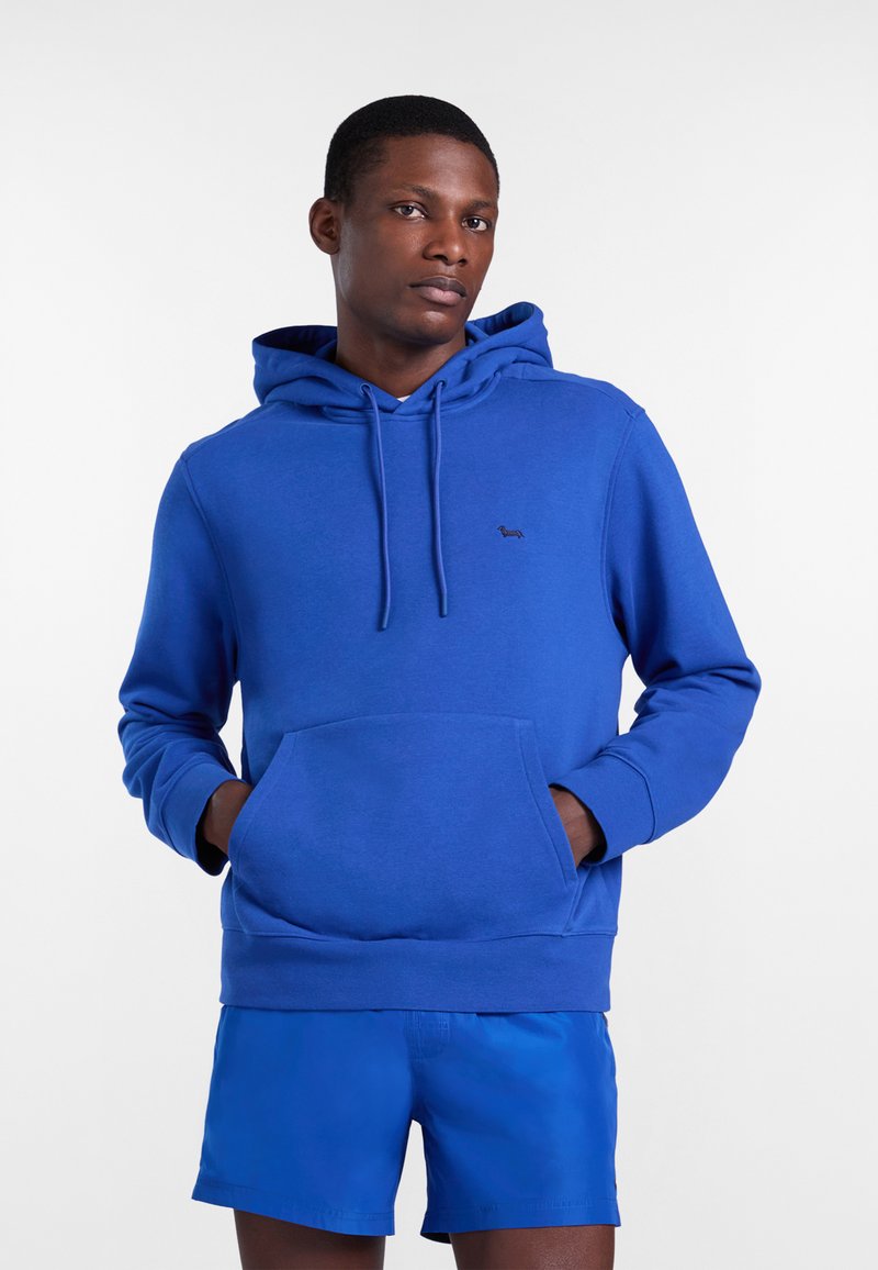 Mężczyzna w niebieskiej bluzie z kapturem i kieszenią kangurka oraz pasujących niebieskich szortach, stojący z rękami w kieszeniach.