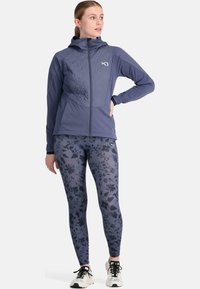 Veste zippée navy avec un devant texturé, assortie à des leggings à motif floral dans des nuances de bleu et de noir. Modèle portant des chaussures de sport.