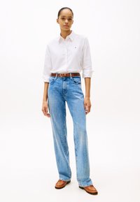 Camisa blanca de botones con mangas arremangadas, jeans de talle alto y pierna ancha en color azul, cinturón marrón y zapatos beige con punta redondeada y detalles de costura.