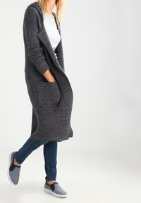 Długi kardigan w kolorze węgielnej szarości z otwartym przodem, dwoma kieszeniami i prążkowanymi rękawami, zestawiony z niebieskimi jeansami i wzorzystymi wsuwanymi butami.