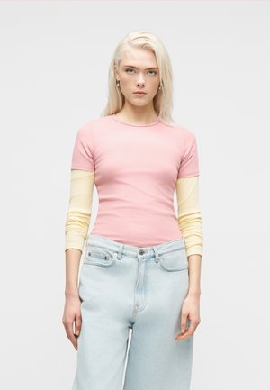 Jeune femme aux cheveux blonds raides portant un haut à manches courtes rose par-dessus un chemisier jaune à manches longues et un jean taille haute bleu clair.