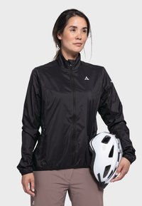 Schwarze Windbreaker-Jacke aus leichtem Material mit hohem Kragen, vorderem Reißverschluss und seitlichen Taschen, kombiniert mit einem weißen Fahrradhelm.